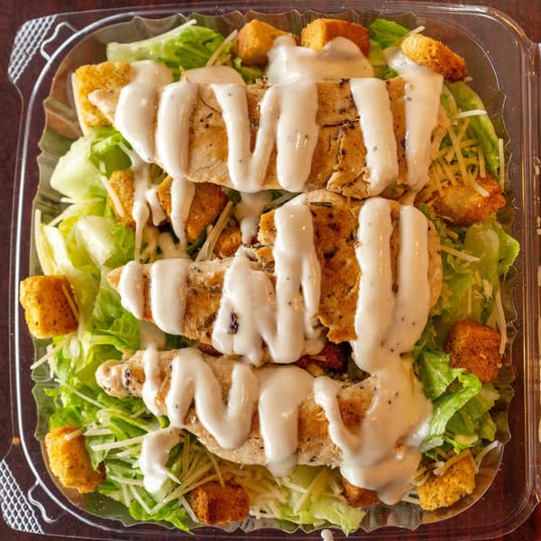 Caesar salad with grilled chicken, croutons, and dressing in a clear plastic container.