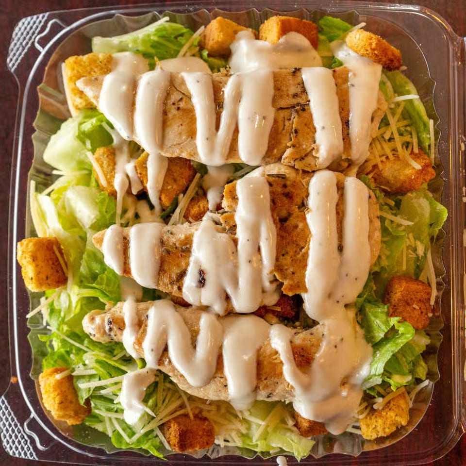 Caesar salad with grilled chicken, croutons, and dressing in a clear plastic container.